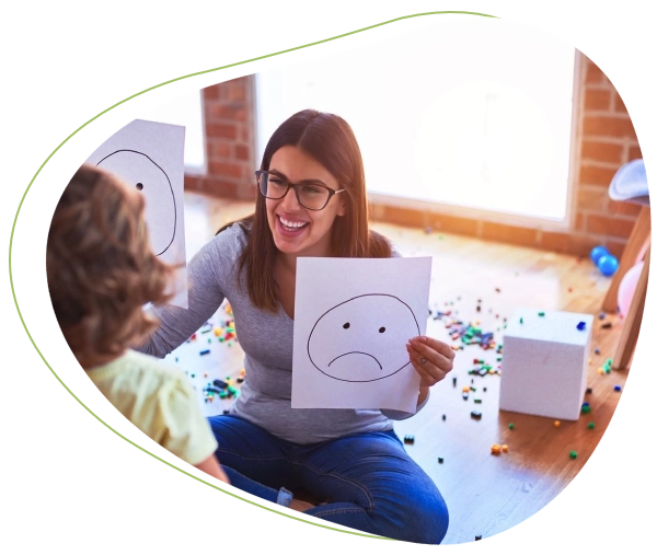 A woman sitting on the floor holding up paper with an unhappy face drawn on it.